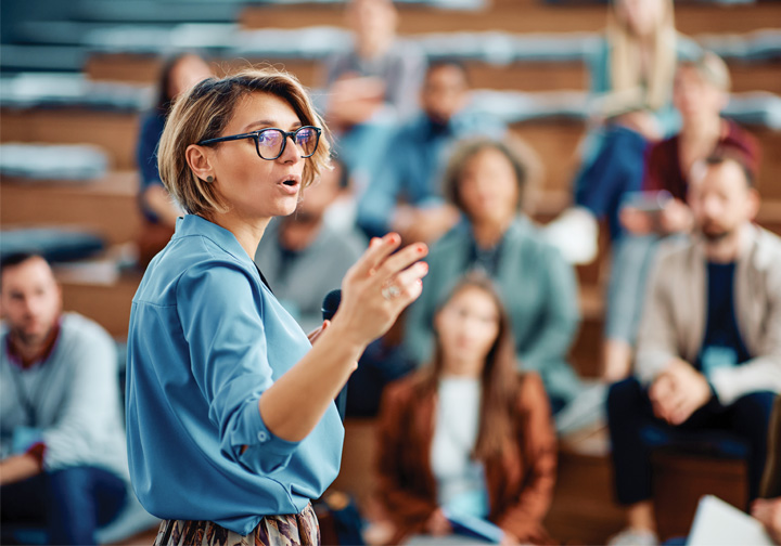A woman in glasses and a blue top gesticulates as she addresses a group of people in the background.
