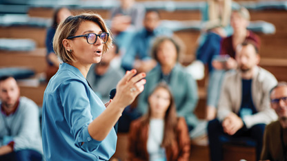 A woman in glasses and a blue top gesticulates as she addresses a group of people in the background.