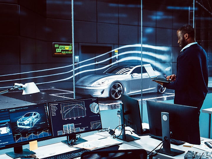 African American male holding a laptop standing in research room with several computers monitoring a grey sports car in wind tunnel