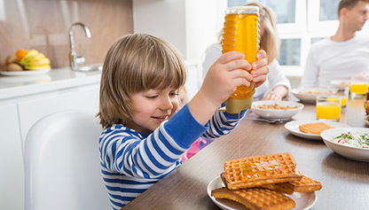 Young blonde boy joyfully squeezing honey onto waffles at the breakfast table with family
