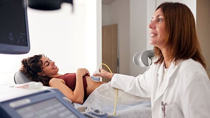 In the middle of the ultrasound exam, the patient looks toward the gynecologist. A female in a lab coat conducts an ultrasound scan on a young woman.