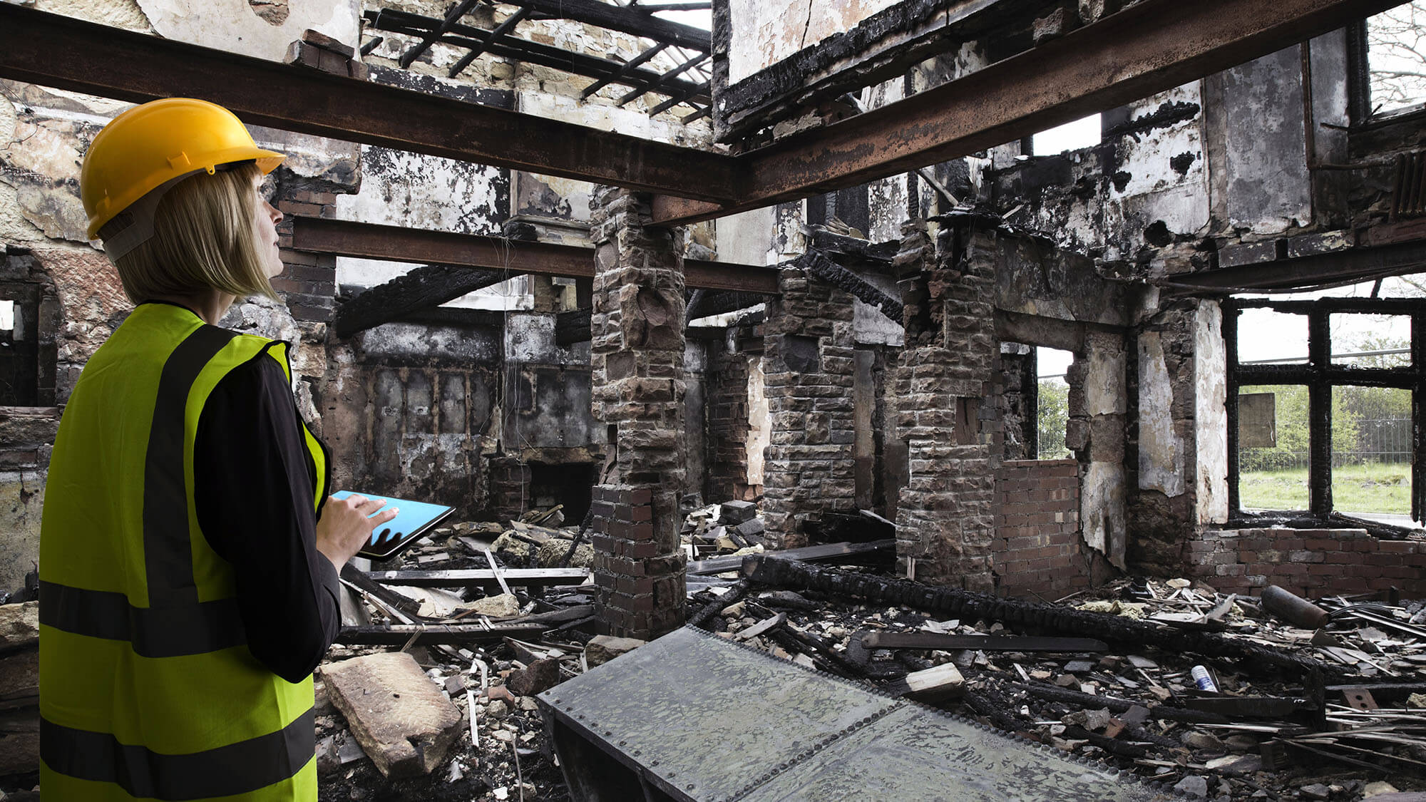 Mature female in a yellow hard hat and caution vest assessing fire damage inside of a property